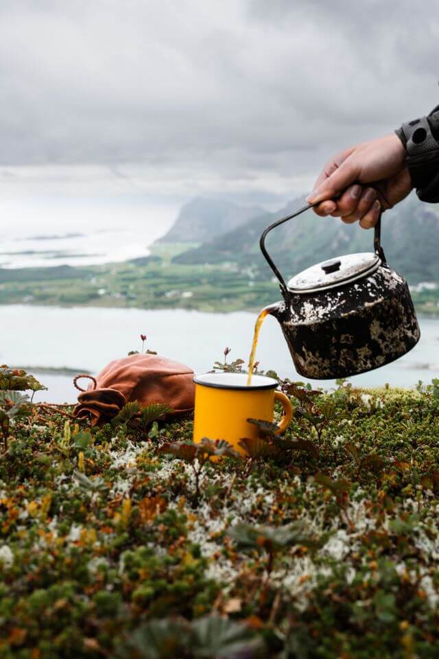 Cut out of a hand pouring tea from an iron kettle into a yellow metal mug which is resting on grass, with a background of a coastline and hills on a cloudy day